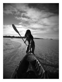 Woman pulling boat while holding oar on shore at beach against sky