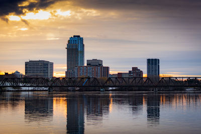 Modern buildings by river against sky during sunset