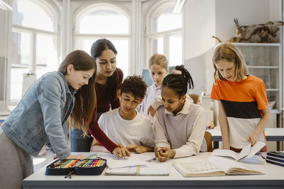 Teacher teaching male and female students with books on desk in classroom