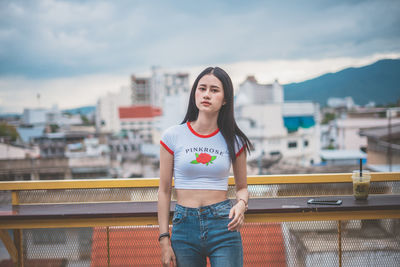 Portrait of young woman standing against sky in city