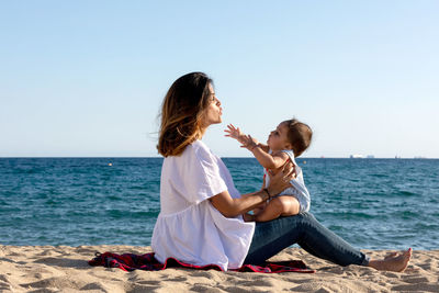 Side view of woman sitting on rock at beach against clear sky