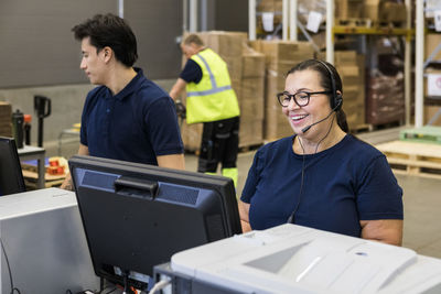 Smiling mature female customer service representative talking through headset while standing by coworker in distribution
