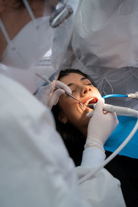 Crop professional dentist in uniform with medical mask drilling tooth of calm woman with help of assistant