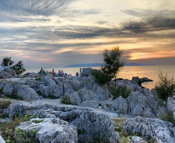 People on rocks by sea against sky during sunset