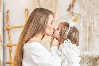 Mom kisses her little daughter sitting on the bed in a bright bedroom.family. ethno-interior design.