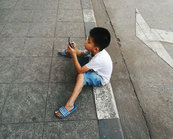 High angle view of boy using phone while sitting on sidewalk