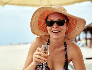 Portrait of woman in sun hat and sunglasses holding champagne at beach