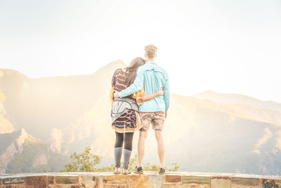 Rear view of couple with arms around standing against mountain sky