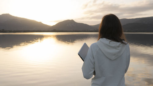 Rear view of woman standing by lake against sky during sunset