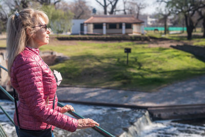 Side view of an adult woman with sunglasses standing in the park holding on handrail with a pond