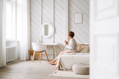 Middle aged plus size woman with brunette curly hair with cup of tea in hands sitting on bed at home