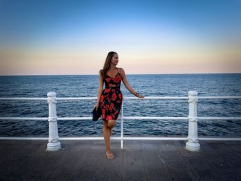 Young woman wearing summer dress leaning on a railing over the ocean at sunset