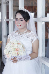 Smiling bride wearing white wedding dress holding bouquet by window
