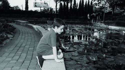 Side view of cute boy looking at water lilies in pond while kneeling on retaining wall at park