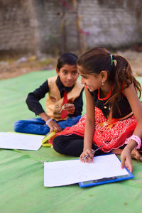 Side view of girl and boy drawing on paper while sitting outdoors