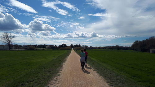 Rear view of friends walking on dirt road amidst green landscape against sky during sunny day