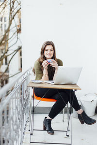 Portrait of smiling young woman with laptop on table having coffee at balcony