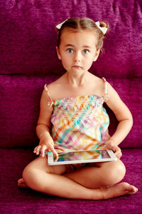Portrait of cute girl using digital tablet sitting on pink sofa at home