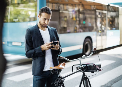 Businessman using mobile phone while standing with bicycle on city street