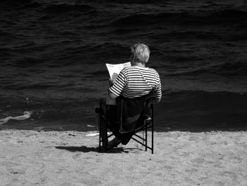 Rear view of man reading newspaper while sitting on champing chair at beach