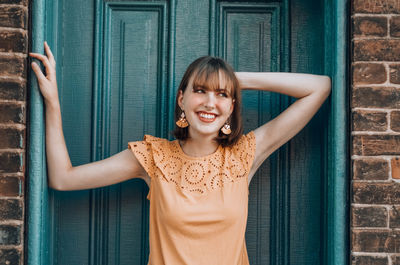 Portrait of smiling young woman standing by door against wall