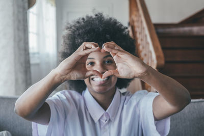 Portrait of smiling woman making heart shape sitting on sofa at home