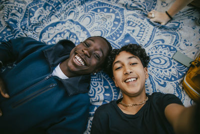 Smiling teenage boy taking selfie with male friend while lying down on picnic blanket