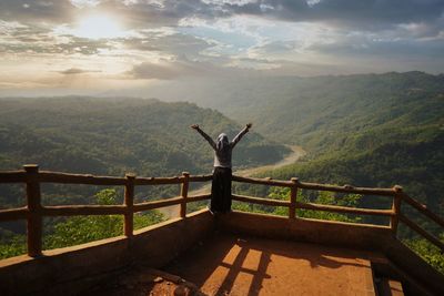 Rear view of man standing on railing against sky during sunset