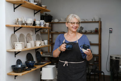 Potter artist showing her work in studio. mature ceramist standing at workshop with clay cups 