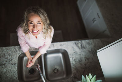 High angle portrait of smiling young woman washing hands in sink