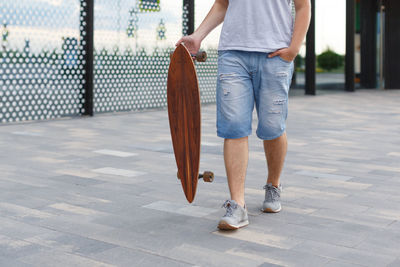 Cropped outdoor picture of man in denim shorts and grey sneakers walking with a longboard