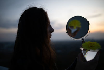 Close-up of woman holding mirror against sky during sunset