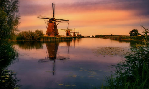 Traditional windmill by lake against sky during sunset