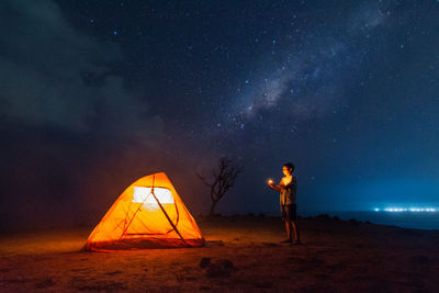 Side view of man standing by illuminated tent at beach during night