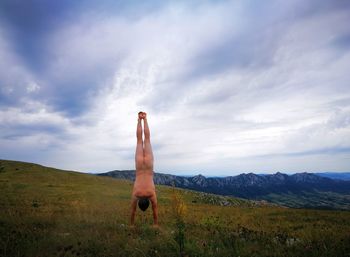 Rear view of naked young man doing handstand on grassy landscape against cloudy sky