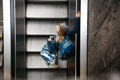 Woman with backpack and travelling bag standing on escalator looking up