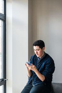 Young businessman using smart phone while sitting on sofa against wall at office