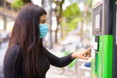 Side view of woman wearing mask using ticket machine