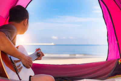 Man sitting in tent and playing guitar on shore of beach