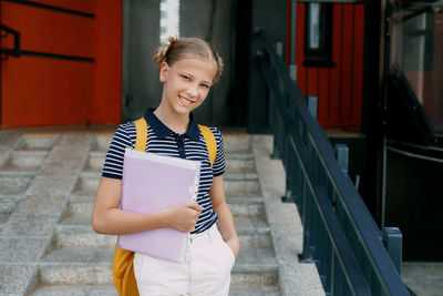 A smiling teenage girl with a backpack and a folder in her hands goes down the stairs