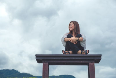 Low angle view of woman sitting on bench against cloudy sky