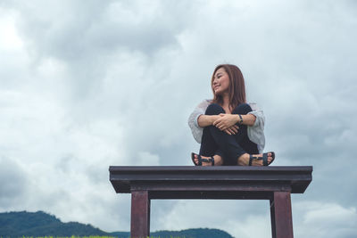Young woman sitting on bench against sky