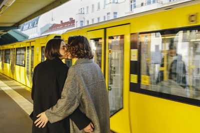 Lesbian couple kissing while standing by yellow train at railroad station