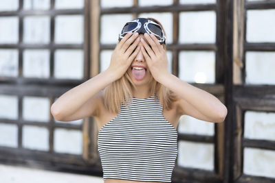 Playful young woman covering eyes with hands while sticking out tongue outdoors