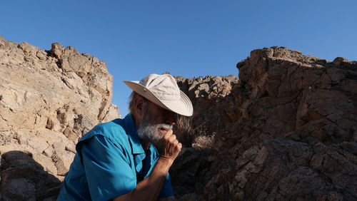 Side view of senior man sitting on rock against clear blue sky