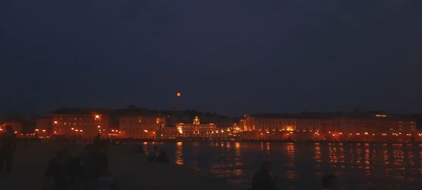 Illuminated city by sea against sky at night