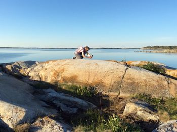 Man photographing while kneeling on rock at beach against sky