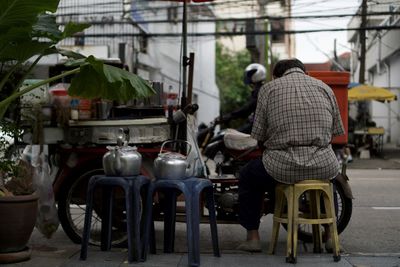 Rear view of old man sitting on food stall on street in bangkok and motorbike running pass