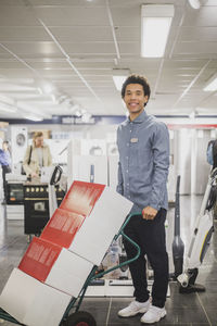 Portrait of smiling male owner with luggage cart standing in electronics store