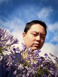 Low angle view of young man holding purple agapanthus lilies against cloudy blue sky.